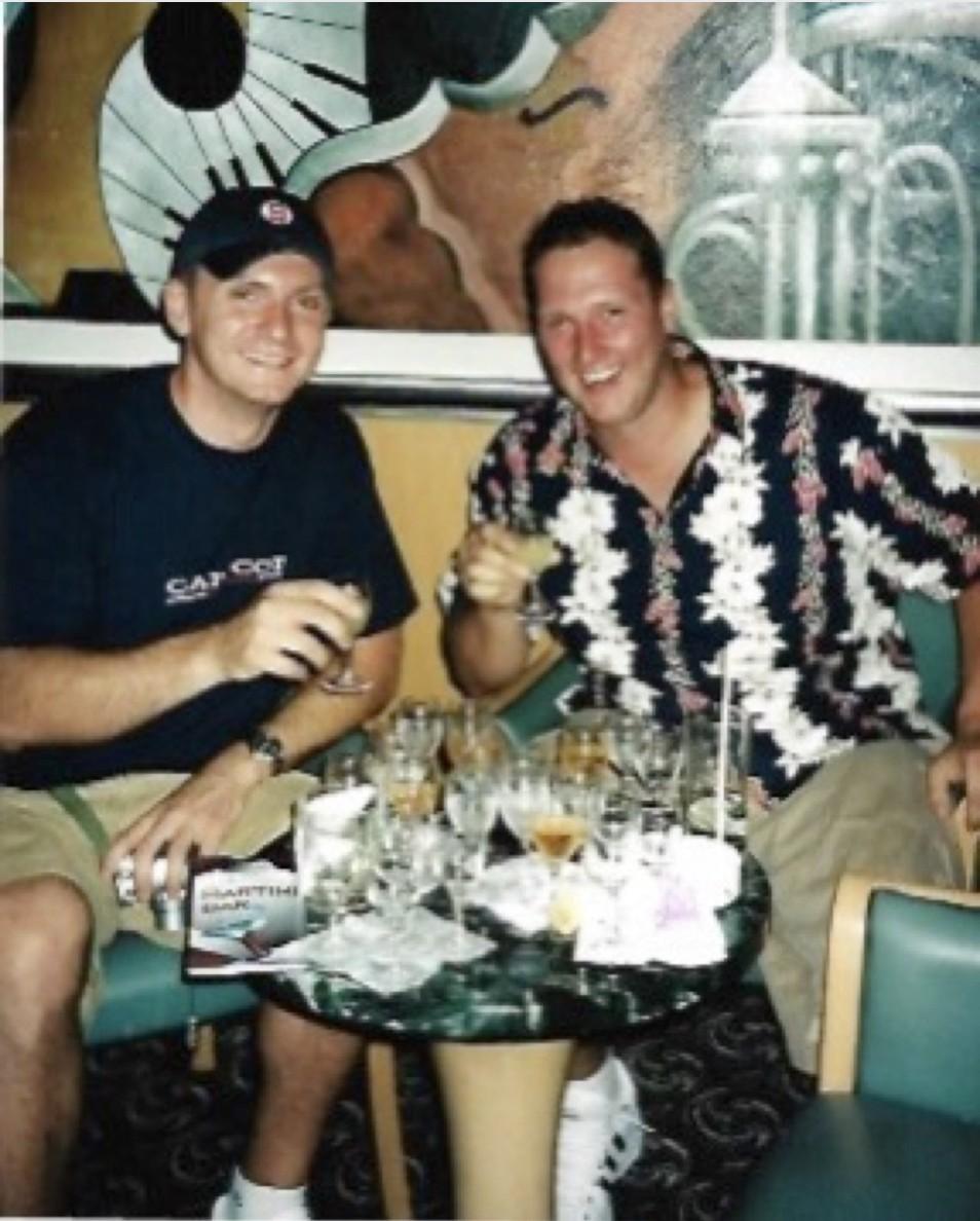 Vintage photo of a young Wayne Partello and his late friend Eddy smiling together at a restaurant table with drinks.