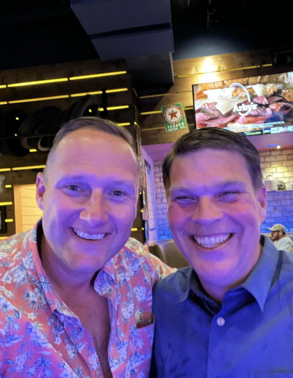 Wayne Partello smiling in a selfie with his mentor Tony at a bar/restaurant, both lit by blue and purple ambient lighting.
