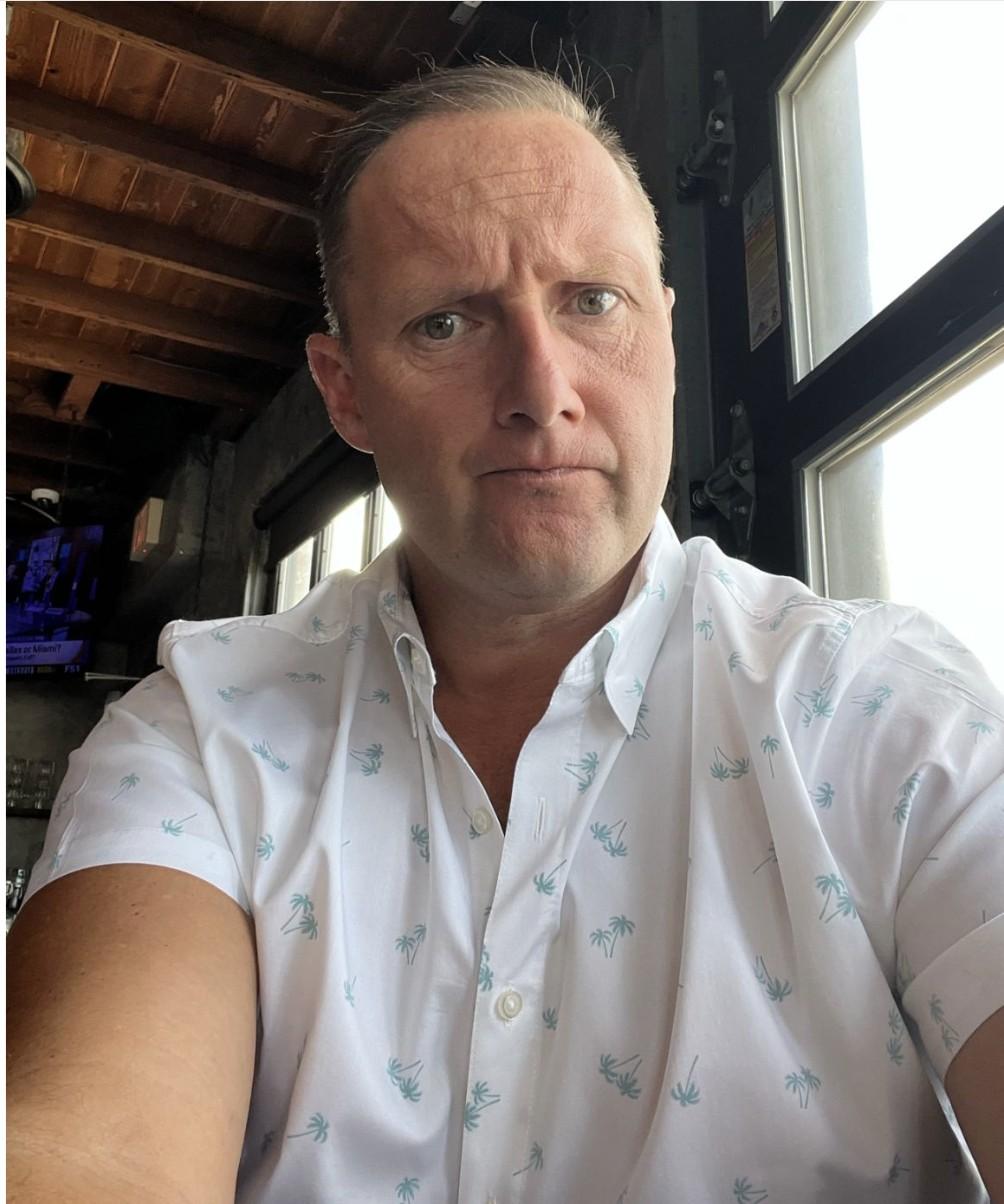 Wayne Partello takes a skeptical-looking selfie wearing a palm tree print shirt.