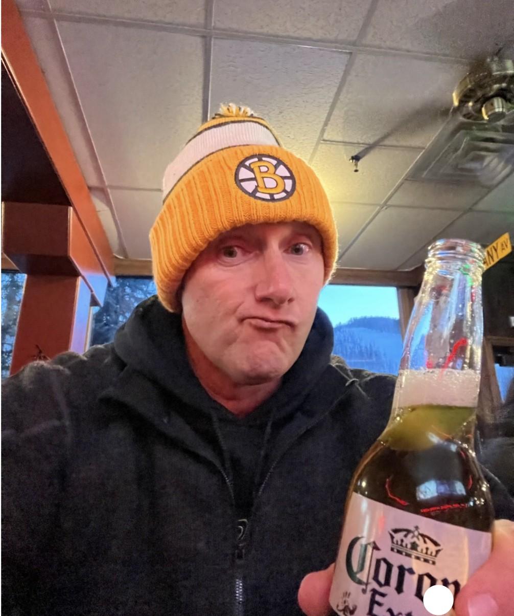 Wayne wearing a yellow Boston Bruins beanie at a ski lodge, holding up a Corona Extra beer.