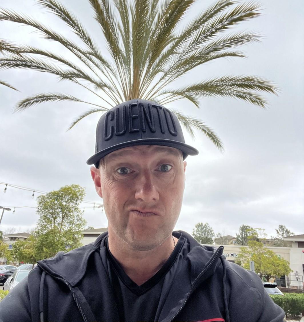 Wayne making a skeptical expression in an outdoor selfie, wearing a CUENTO cap and hoodie under a palm tree.