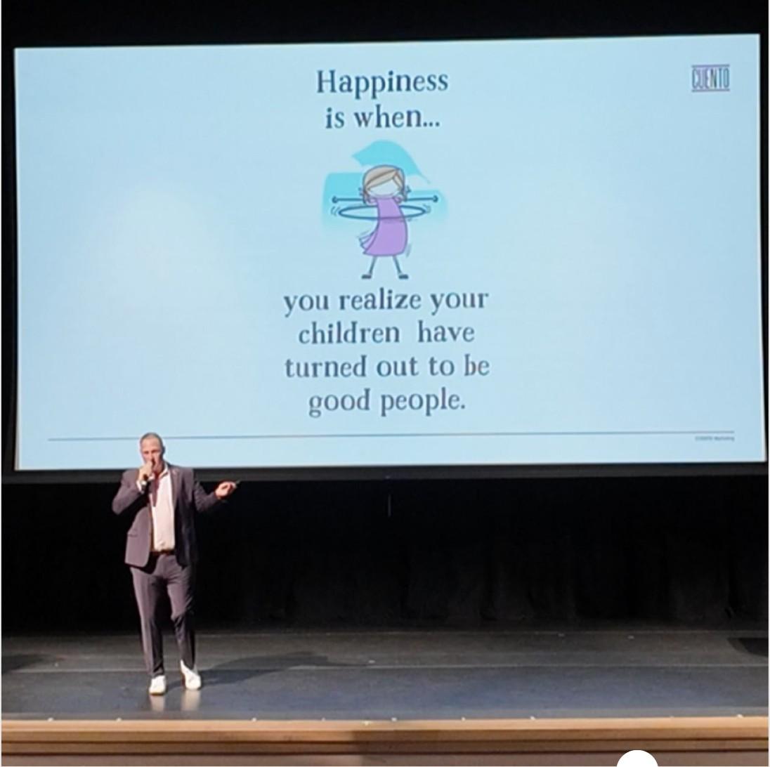 Wayne speaking on a school auditorium stage with a microphone, in front of a large CUENTO-branded presentation slide reading 'Happiness is when... you realize your children have turned out to be good people.'