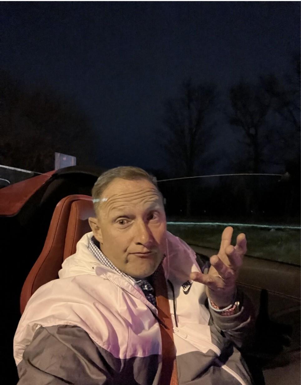 Wayne sitting in a convertible car at night, making an expressive hand gesture toward the camera.