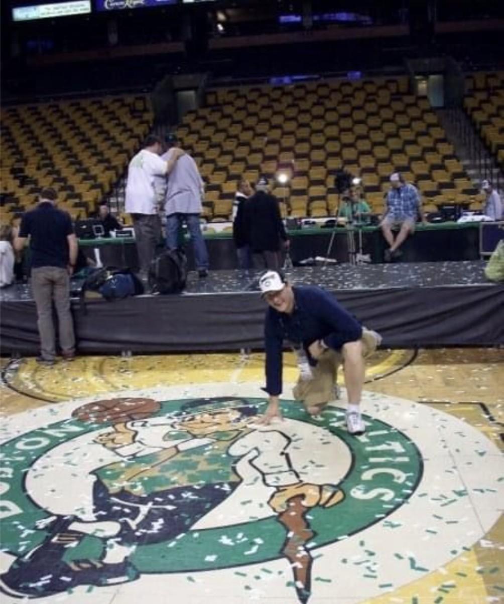 A young Wayne kneeling on the Boston Celtics parquet floor logo amid confetti after the 2008 NBA championship celebration, with the empty arena in the background.