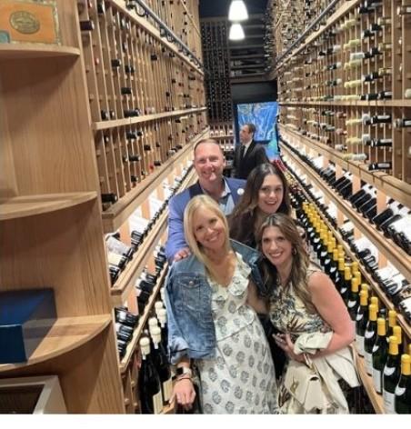 Wayne and three friends posing together in a wine cellar between rows of shelved bottles.