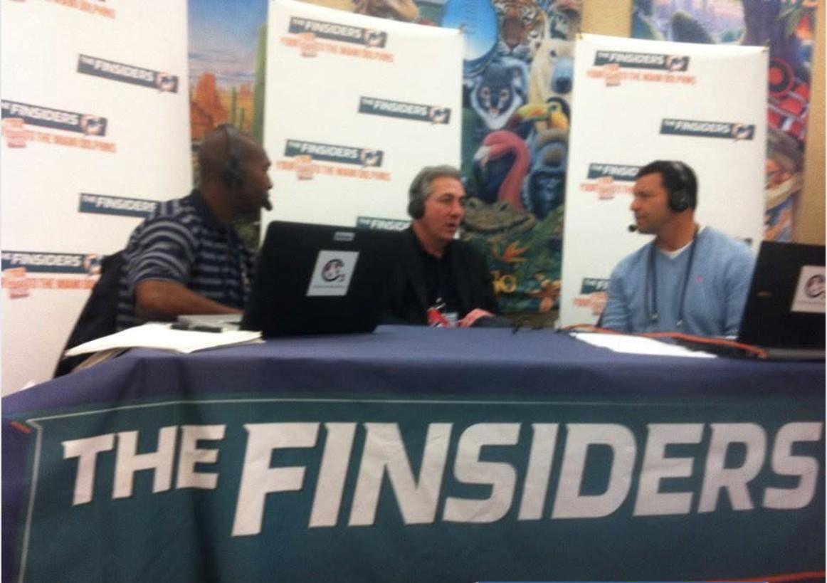 Three broadcasters wearing headsets at a table branded 'The Finsiders' with backdrop banners reading 'The Finsiders: Your Guide to the Miami Dolphins'