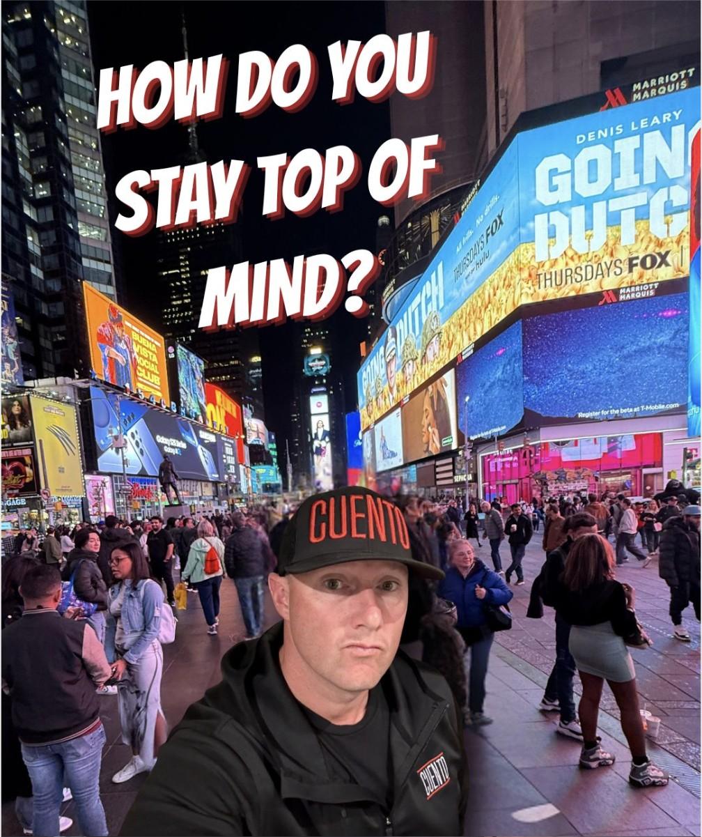 Wayne wearing a CUENTO cap and jacket, taking a selfie in Times Square at night, captioned 'How Do You Stay Top of Mind?'